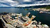 Et stort verft med kaier, skip og industribygg ved en fjord, med fjell og by i bakgrunnen.
