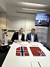 To menn signerer avtaler ved et bord med norske og britiske flagg og en GT Wings-modell.