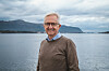 Eldre mann med briller står foran en fjord med fjell og overskyet himmel.