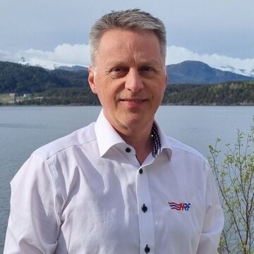Bjarne Rygg i hvit skjorte står ved fjorden med fjell og skog i bakgrunnen.