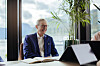 En mann i blå blazer sitter ved et bord i et lyst kontor med planter, notatbok og laptop.