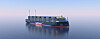 Et blått containerskip seiler på stille hav under klar himmel.