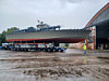 En grå torpedobåt står på en stor transportvogn foran en industrihall på våt asfalt.