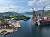 Industrifartøy med kraner i en havn, med fjord, skog og fjell i bakgrunnen.