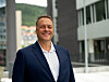 Smilende mann i mørk dress står utendørs foran moderne kontorbygninger.