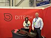 To personer ved en rød DITECH-messestand med en demonstrasjonsenhet på et bord.