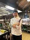 Kokk i hvit uniform står med armene i kors i en skipsbysse med gryter og kjøkkenutstyr.