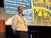 Foredragsholder foran en storskjerm med norsk tekst og industribilder.